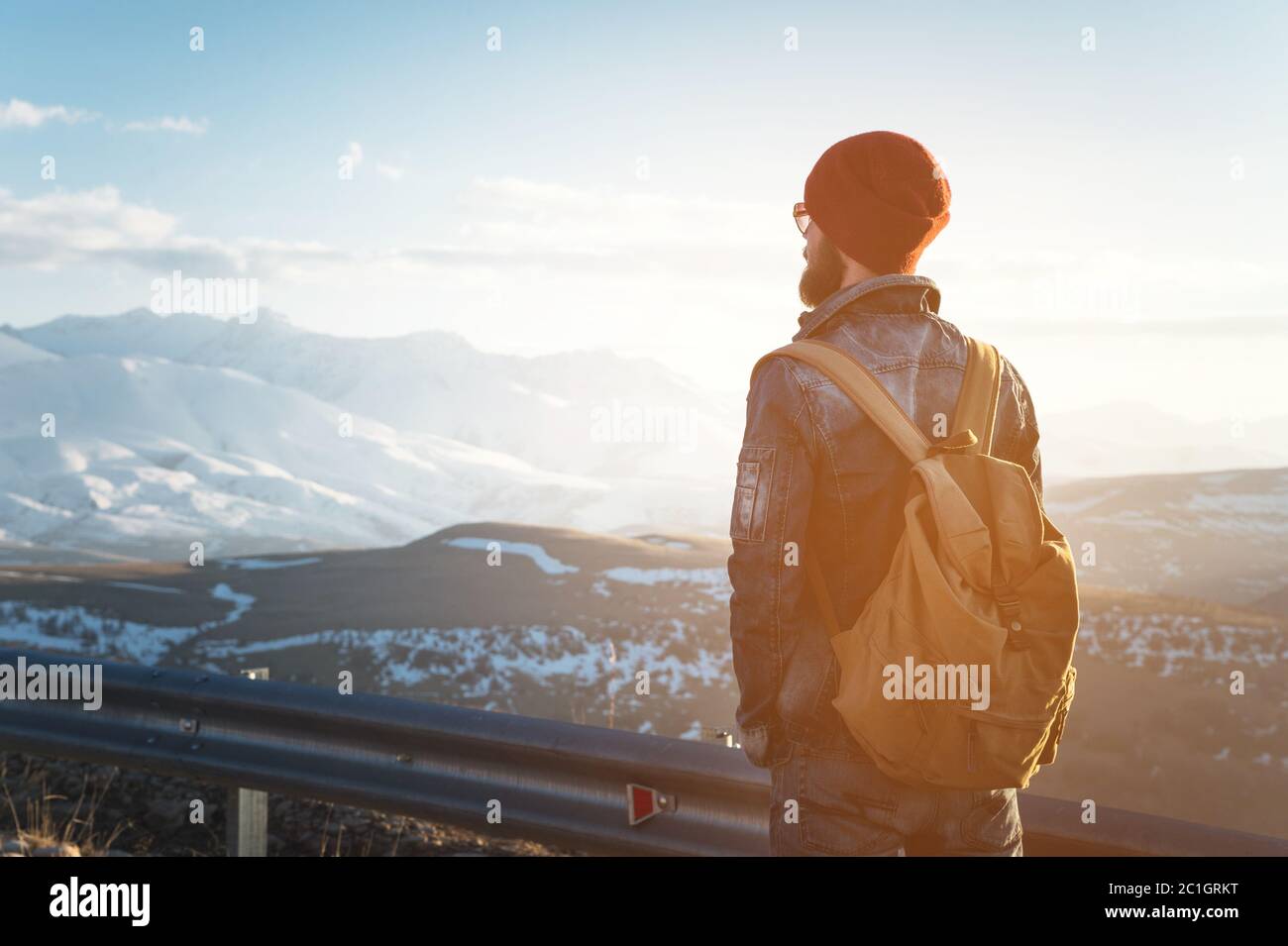 Bearded tourist hipster man in sunglasses with a backpack stand back on a roadside bump and watching the sunset against the back Stock Photo