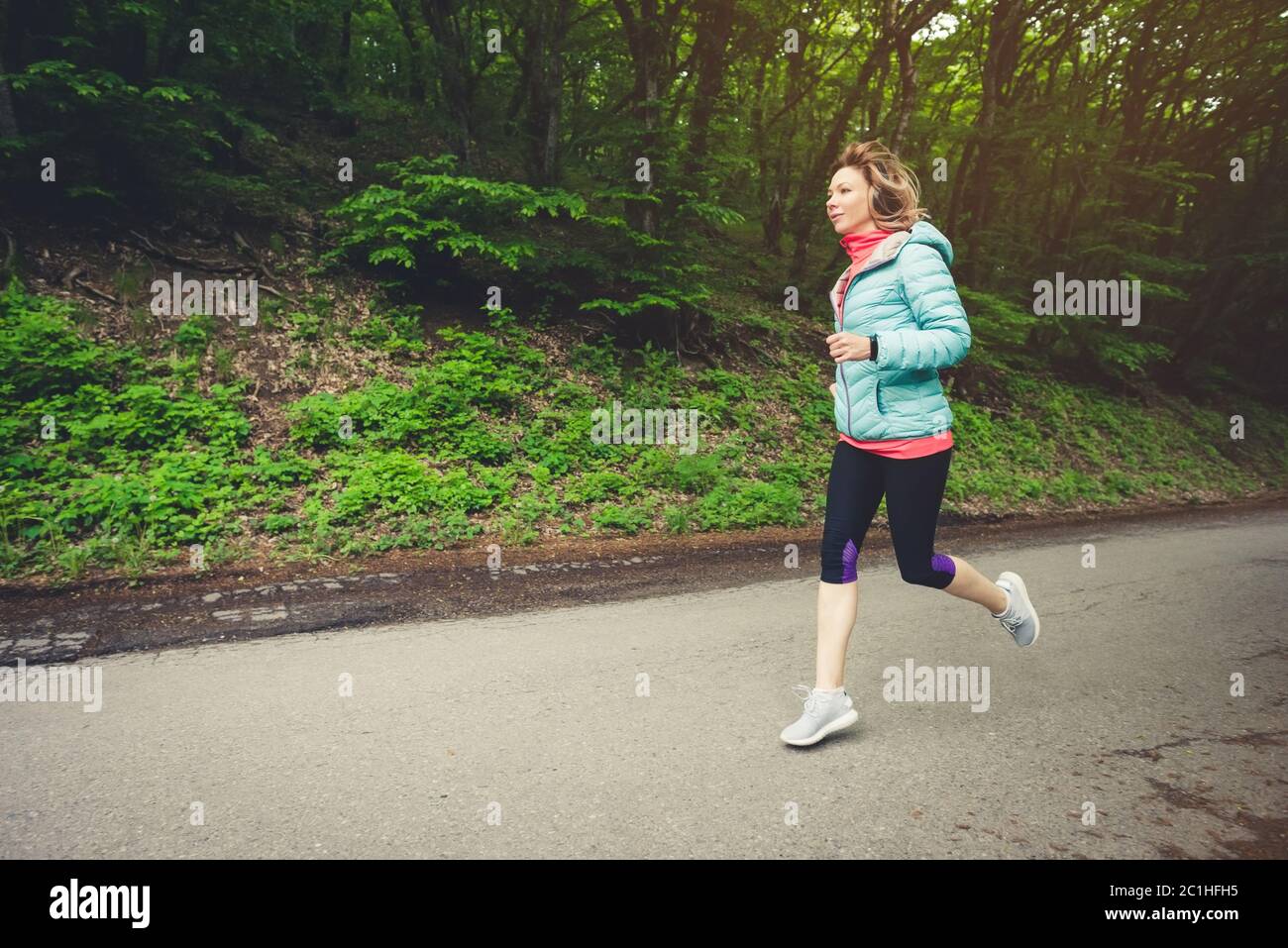 Young fitness blonde woman in headphones running at morning caucasian forest trail in sun light Stock Photo