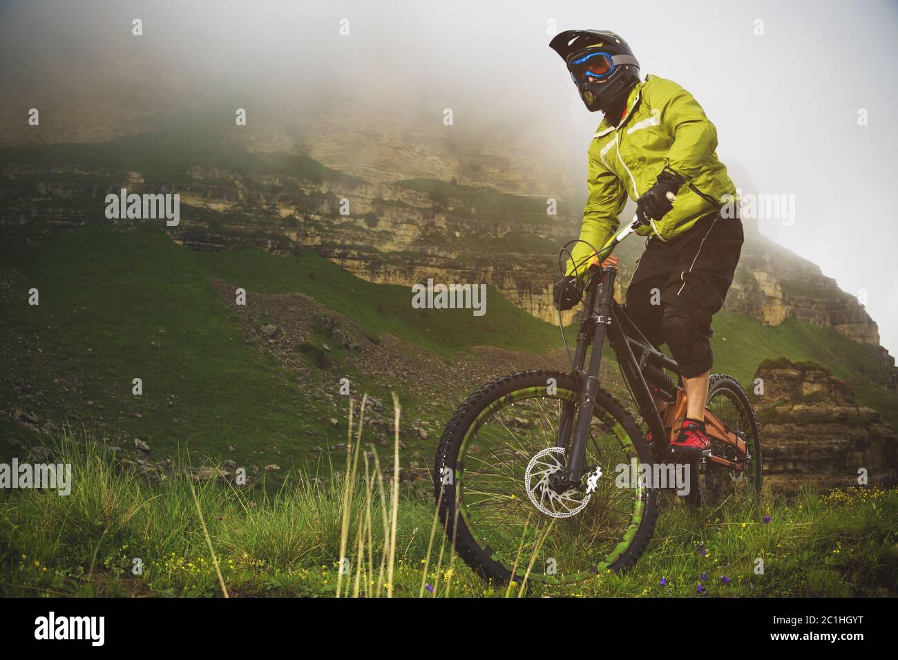 An adult mtb cyclist on a mountain bike at the foot of a cliff surrounded by green grass. Low clouds. North Caucasus. Russia Stock Photo