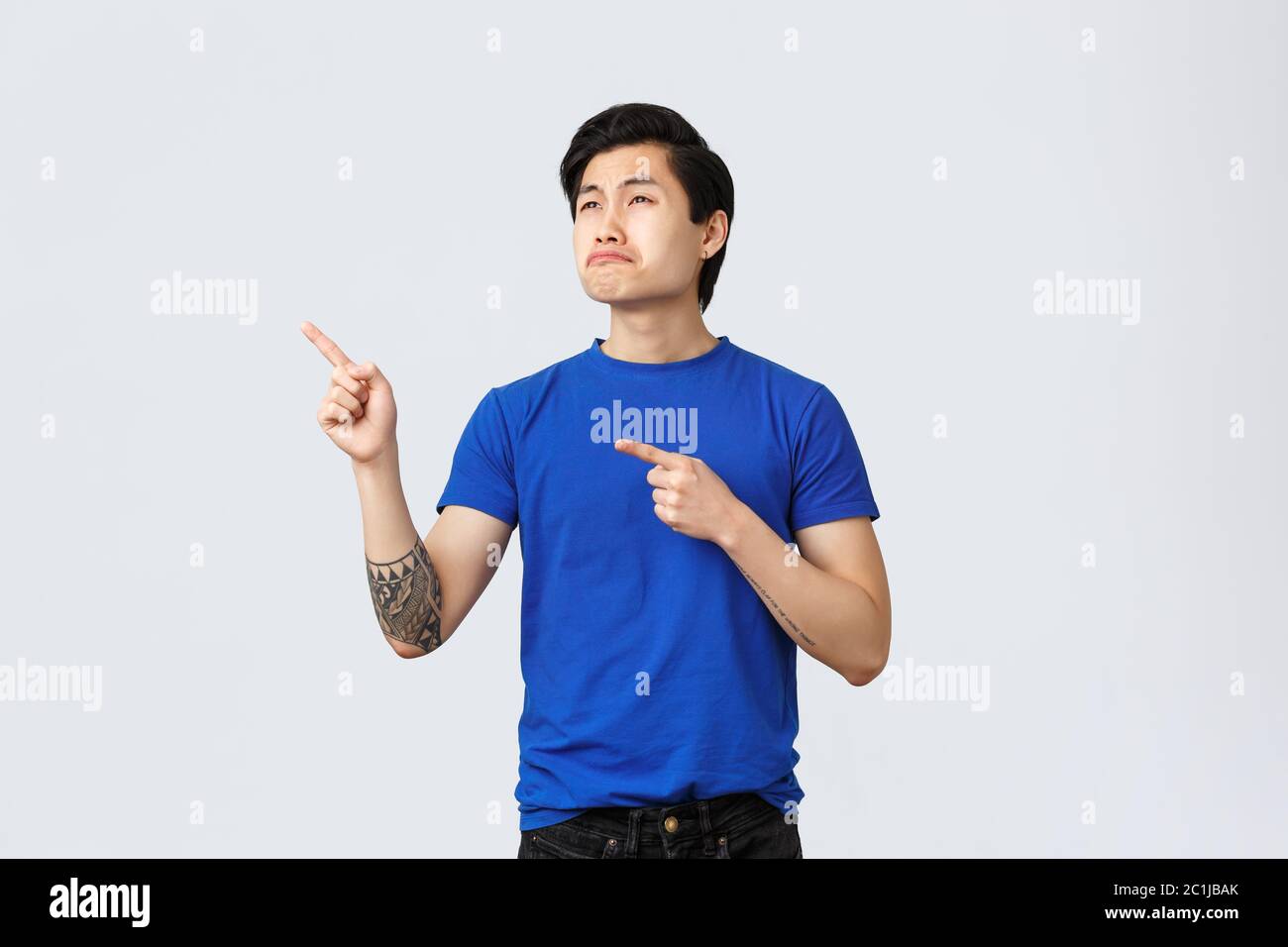 Disappointed and upset cute asian guy, man in blue t-shirt sobbing and looking at promo with jealousy or regret, grimacing pouting, whining at banner Stock Photo
