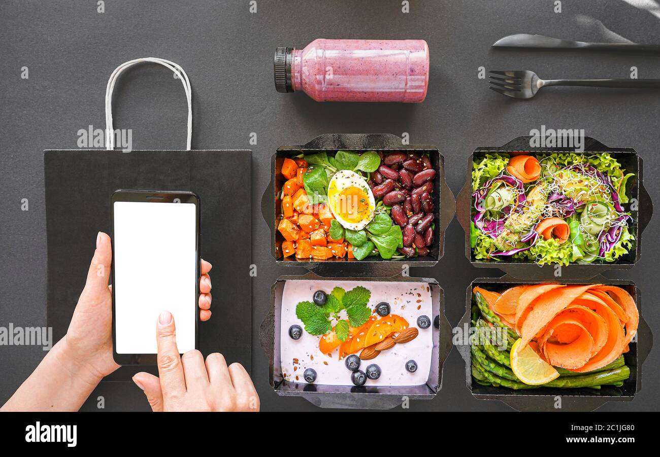 Female hand holding phone mock up screen over vegan healthy food delivery boxes. Stock Photo