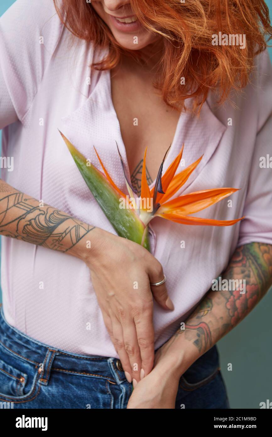 Beautiful girl with red hair and a tattoo is holding a flower strelitzia on a blue background Stock Photo