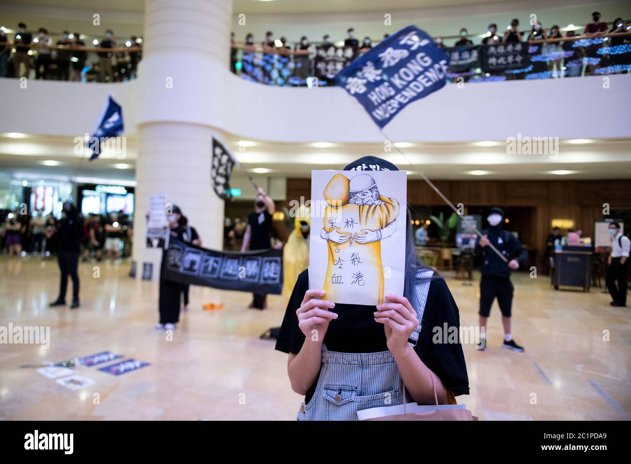 HONG KONG,HONG KONG SAR,CHINA: JUNE 15th 2020. A protester holds up an artwork showing the “raincoat man” in Pacific Place Mall in Admiralty Hong Kong while other wave pro independence signs and sign “Glory to Hong Kong’; the protest anthem. They are there to commemorate the first democracy protesters death on one year ago. Marco Leung fell to his death from scaffolding outside the building on 15th June 2019. He became known as the raincoat man.  Alamy Stock Image/Jayne Russell Stock Photo