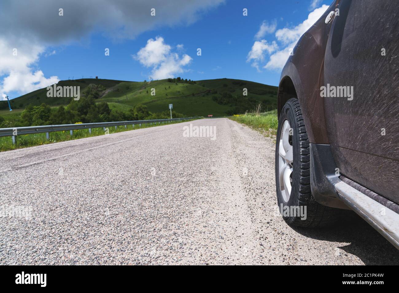 Close-up of a SUV wheel on an asphalt road outside the city against the green hills of blue sky and clouds Stock Photo