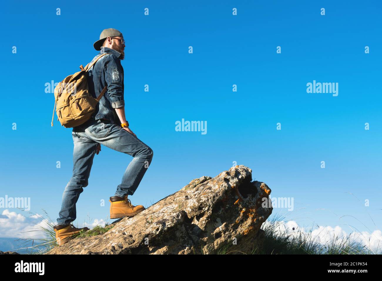 Stylish bearded male traveler in sunglasses and a cap with a backpack in a denim suit and yellow shoes stands on a large stone a Stock Photo