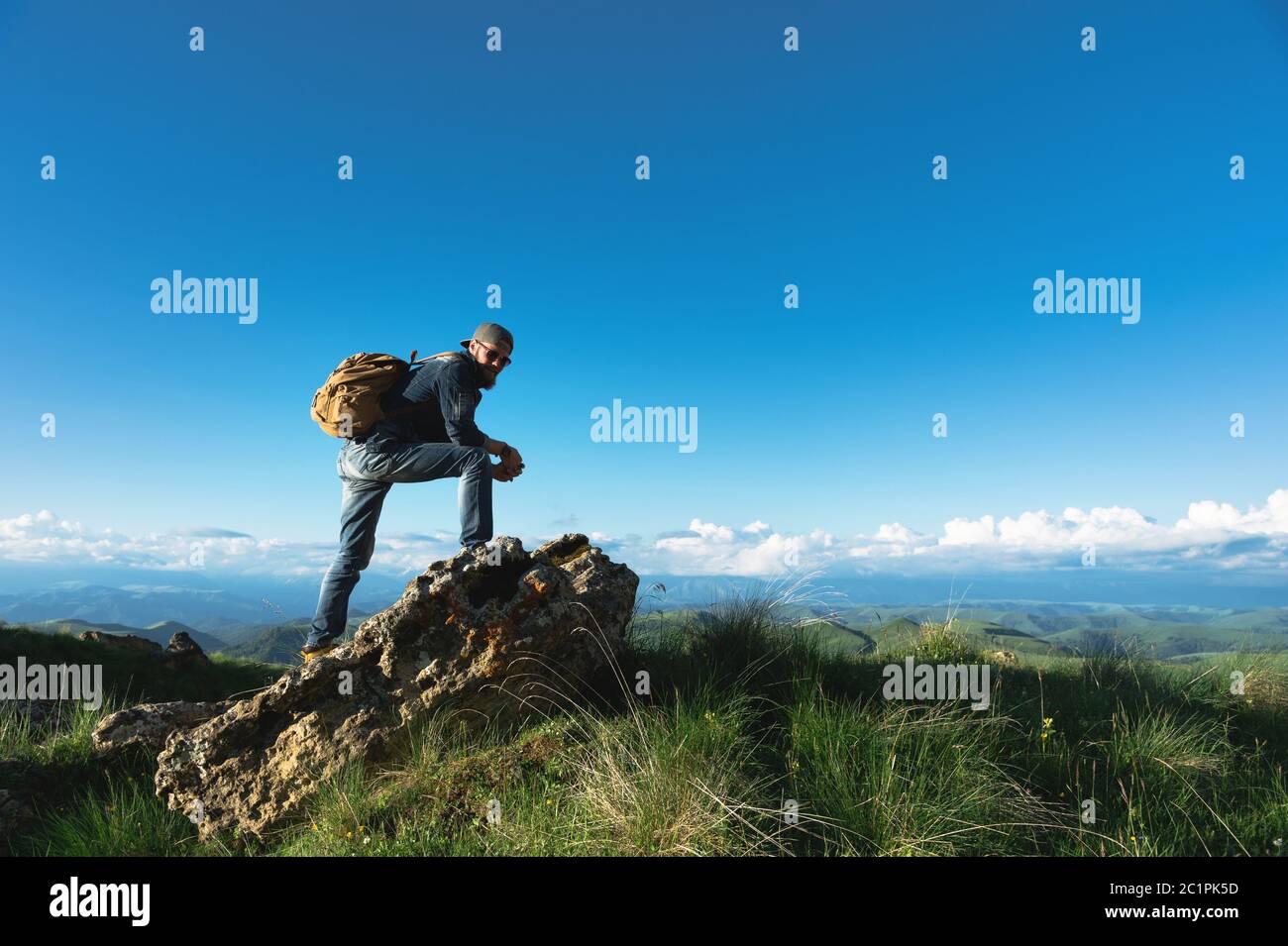 Stylish bearded male traveler in sunglasses and a cap with a backpack in a denim suit and yellow shoes stands on a large stone a Stock Photo