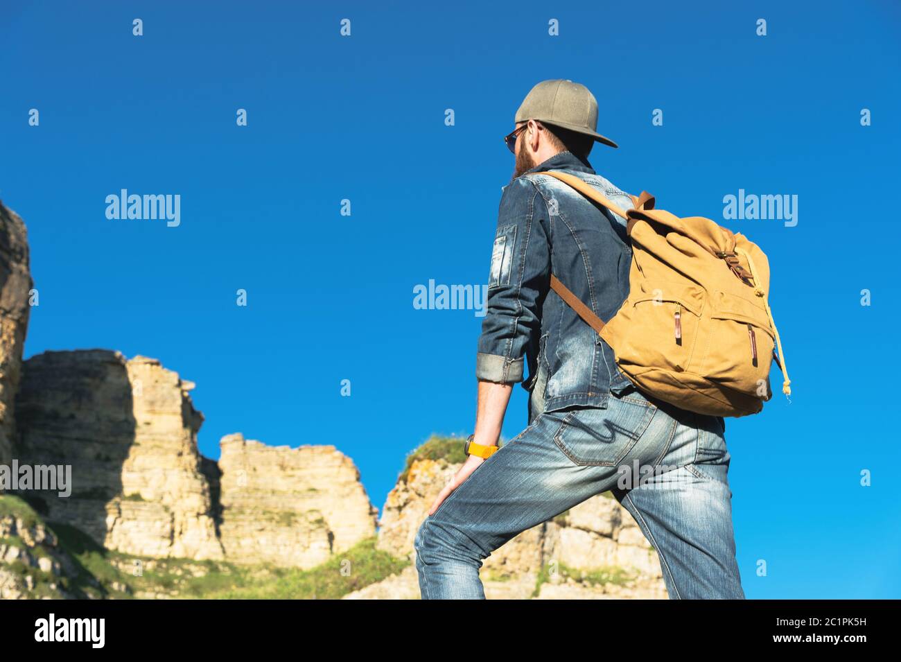 Stylish bearded male traveler in sunglasses and a cap with a backpack in a denim suit and yellow shoes stands with his back on a Stock Photo