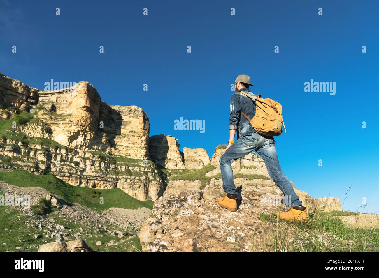 Stylish bearded male traveler in sunglasses and a cap with a backpack in a denim suit and yellow shoes stands with his back on a Stock Photo