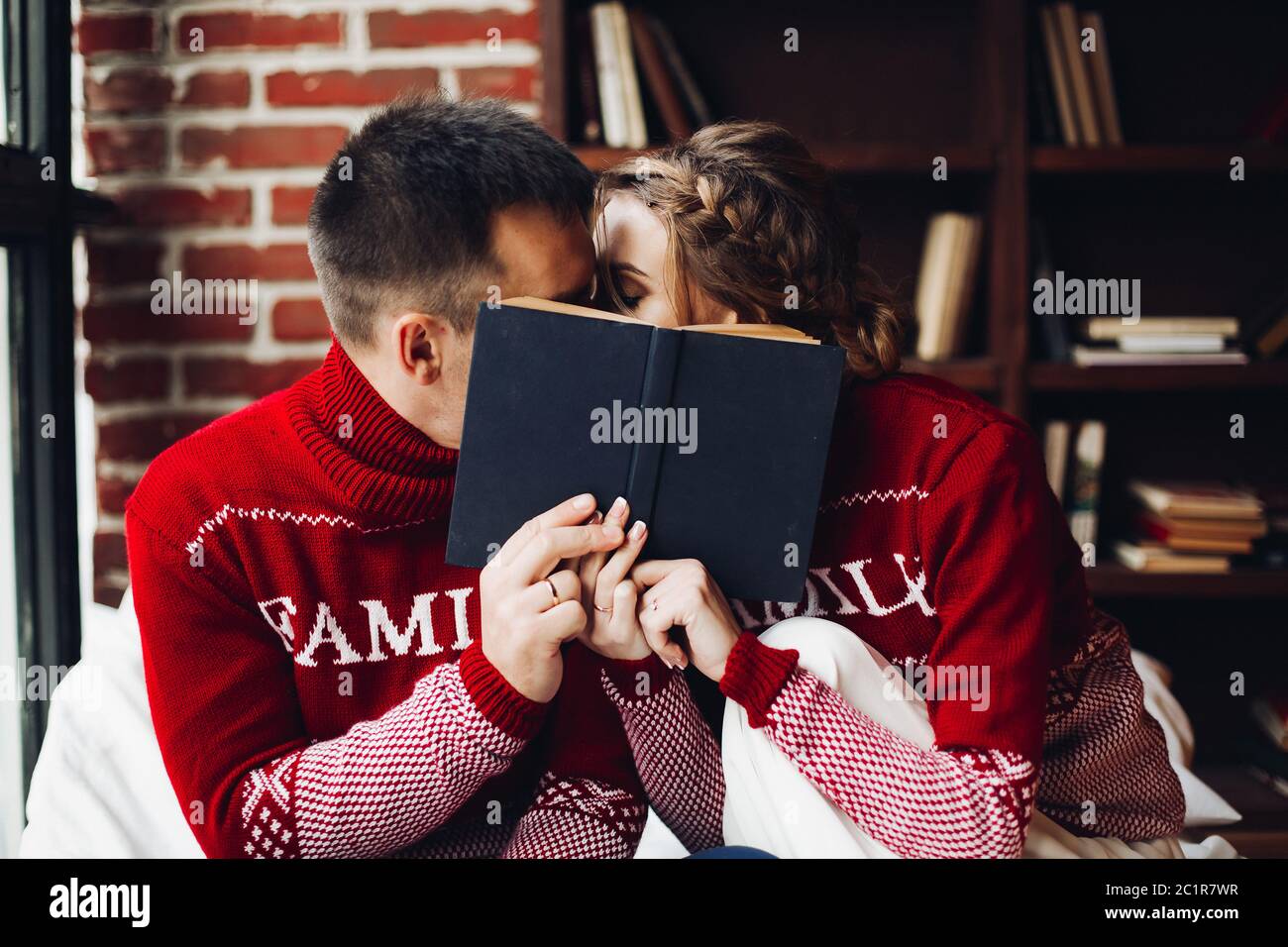 Loving couple in winter jumpers kissing hiding behind the book. Stock Photo