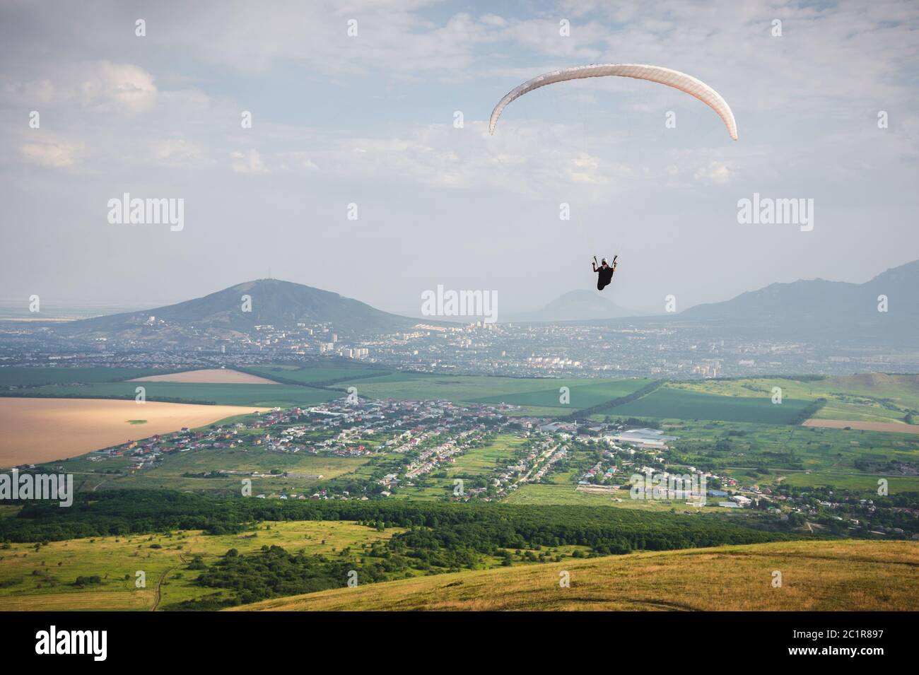 Professional paraglider in a cocoon suit flies high above the ground against the sky and fields with mountains Stock Photo