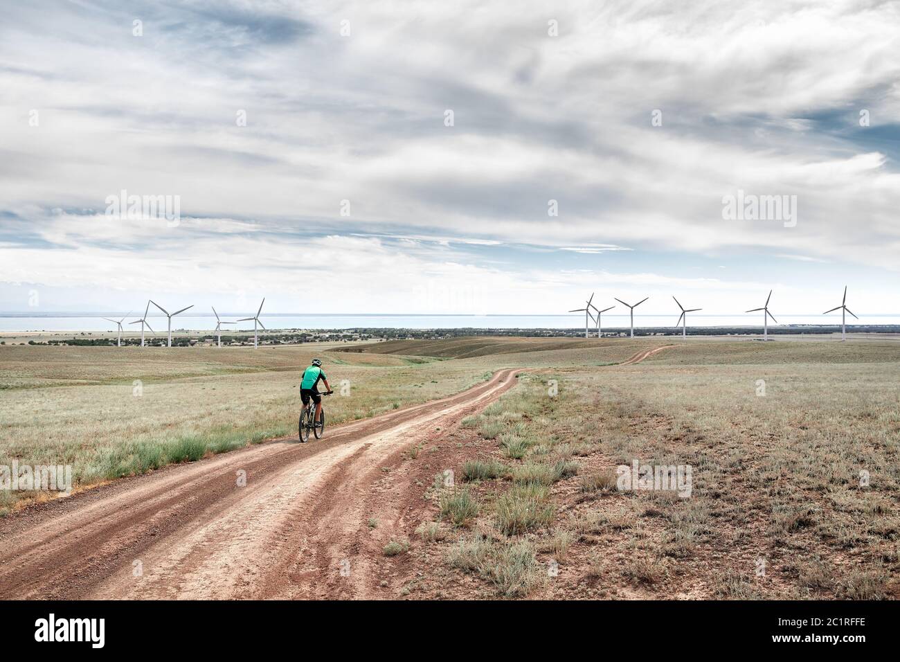 Man on mountain bike rides on the road to the wind turbines farm near the lake Against blue cloudy sky. Ecological and zero waste lifestyle concepts. Stock Photo