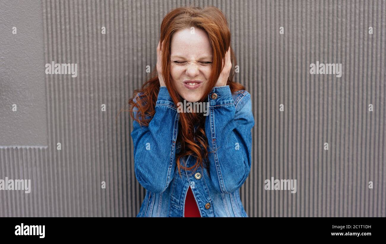 Funny young woman covering her ears with her hands and pulling a funny face with closed eyes in front of a grey textured wall with copy space Stock Photo