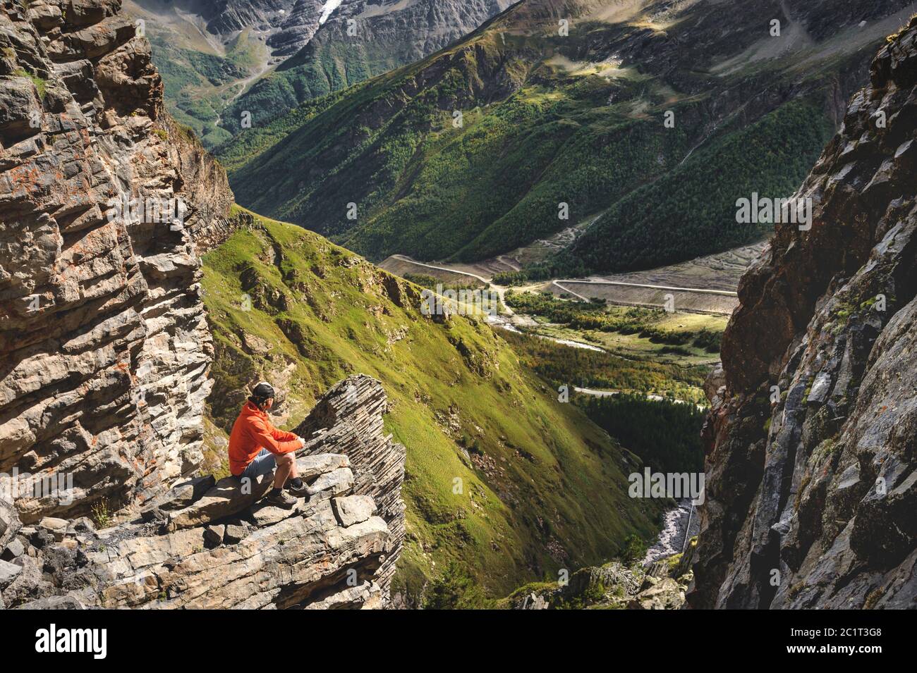 Portrait of a male hiker in shorts and a cap from the back is standing on high rocks at the edge of a cliff against the backgrou Stock Photo