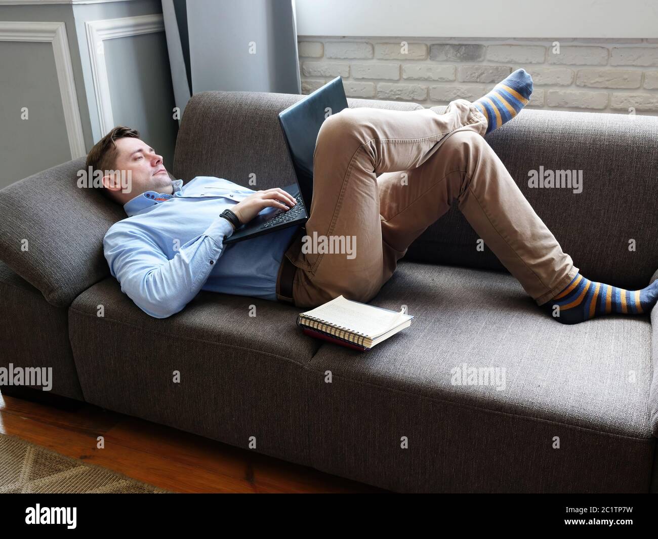 A man lies at home on the couch and works on a laptop. Remote work concept. Stock Photo