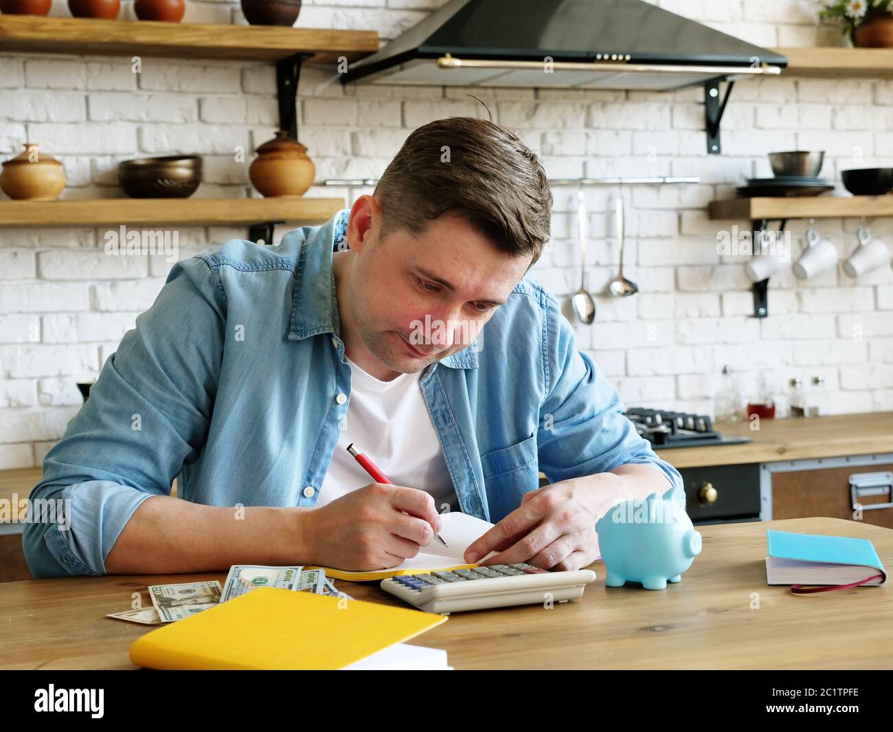 A young man at home calculates the monthly budget. Calculator and piggy bank. Stock Photo