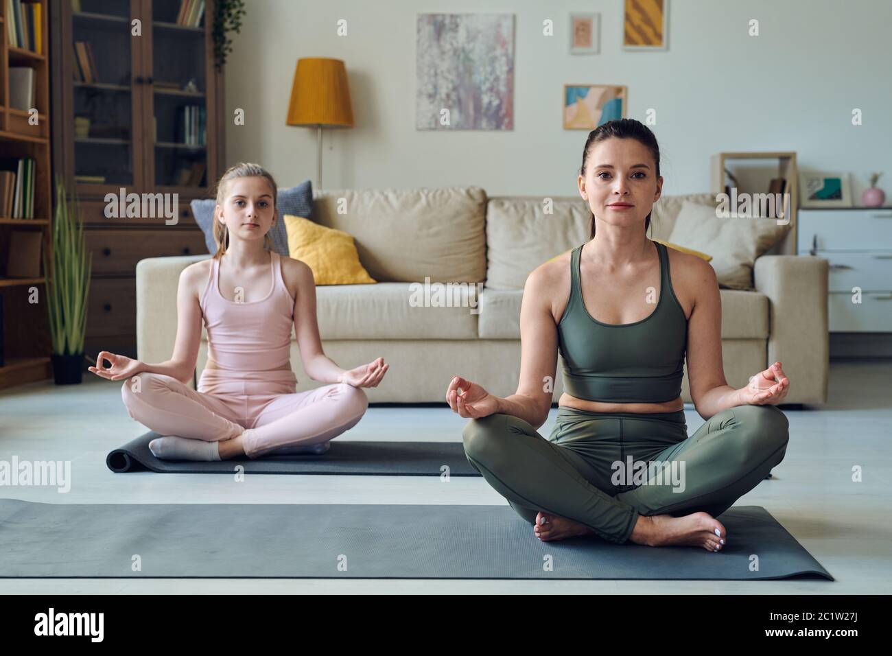 Portrait of sporty mother and teenage daughter sitting with crossed legs while practicing meditation together at home Stock Photo