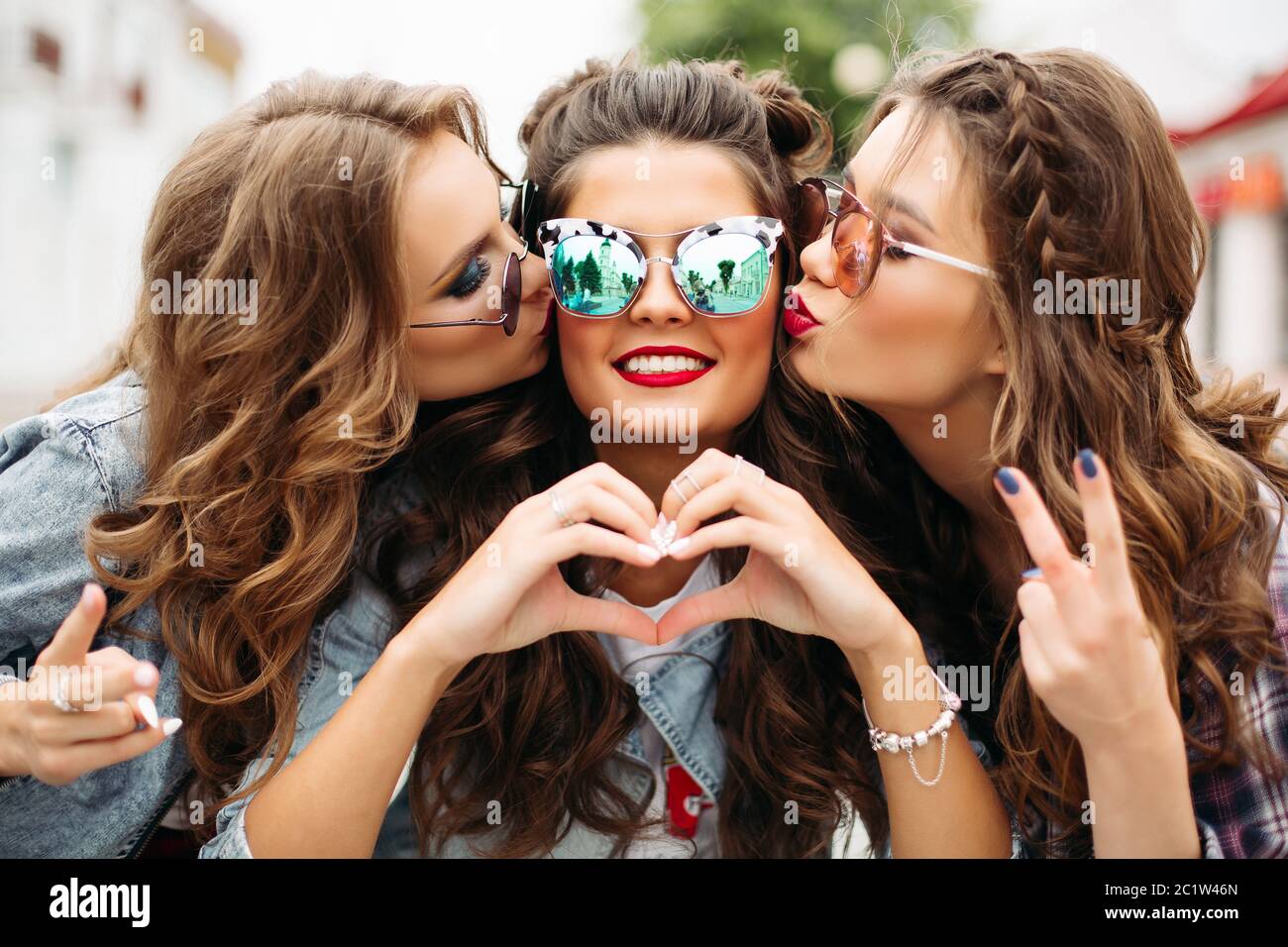 Gorgeous teenagers in sunglasses kissing their friend smiling at camera with heart gesture. Stock Photo