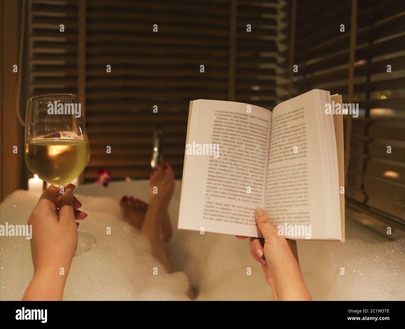 Young woman reading the book and drinking white wine in bath with foam and candles Stock Photo