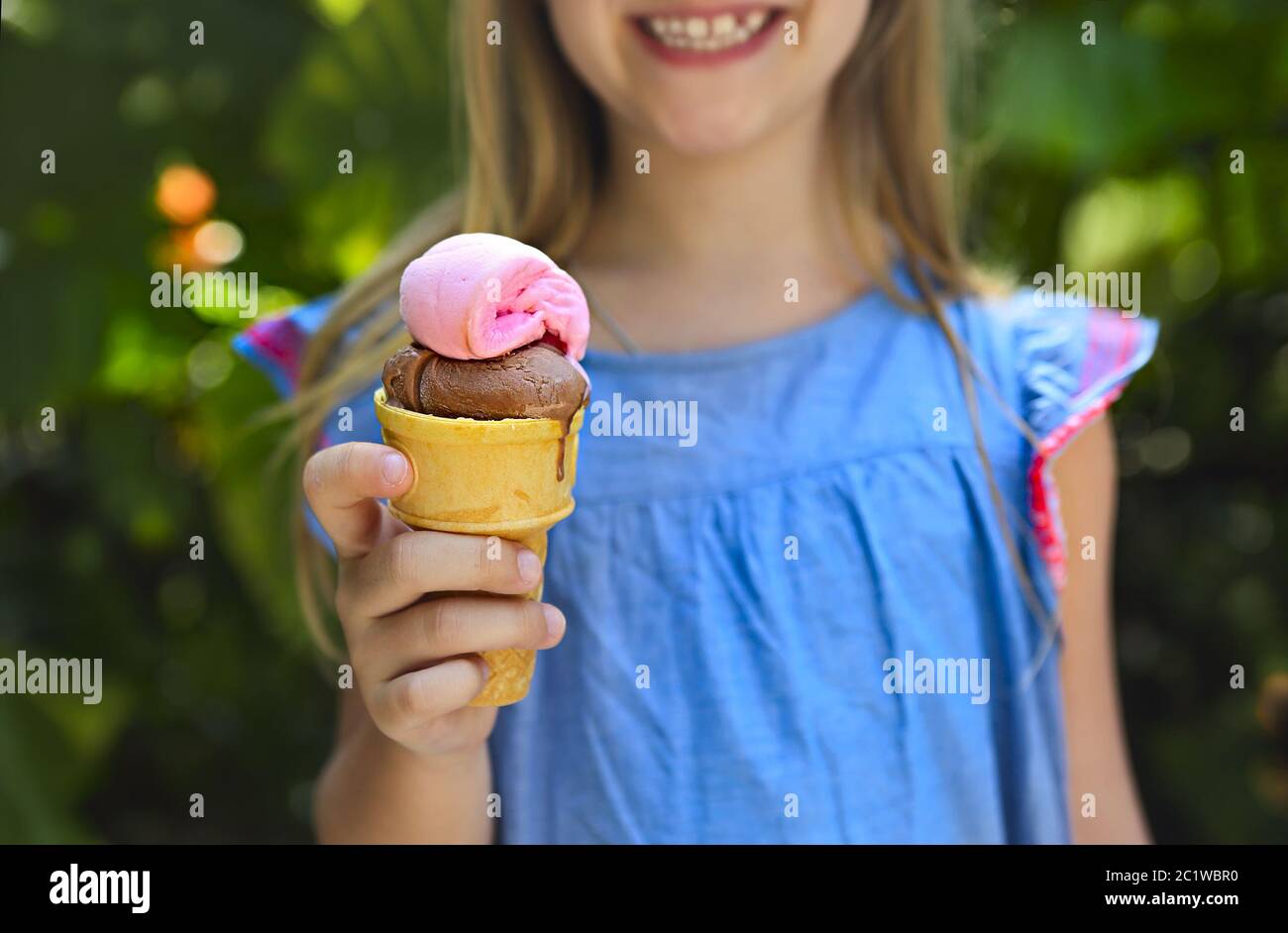 Cute little girl with funny expression holding ice cream cone outside against bright nature background Stock Photo