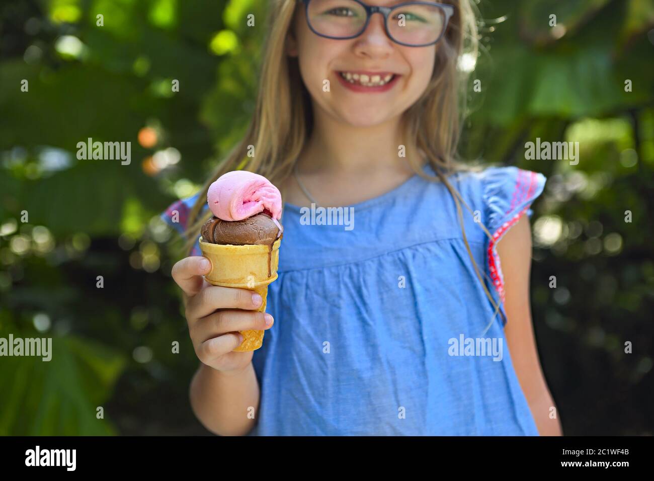 Cute little girl with funny expression holding ice cream cone outside against bright nature background Stock Photo