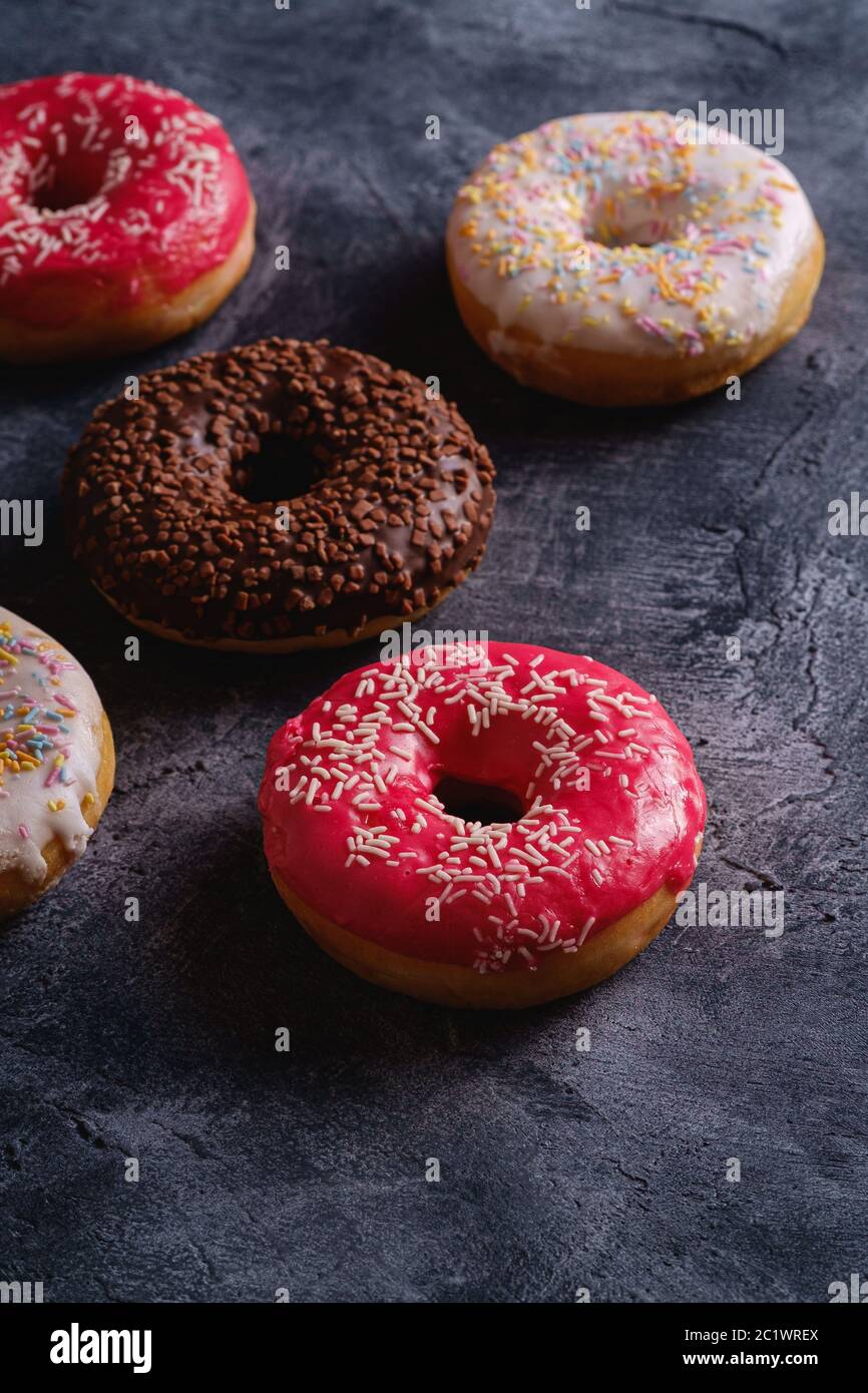 Chocolate, pink and vanilla donuts with sprinkles, sweet glazed dessert food on dark concrete textured background, angle view Stock Photo