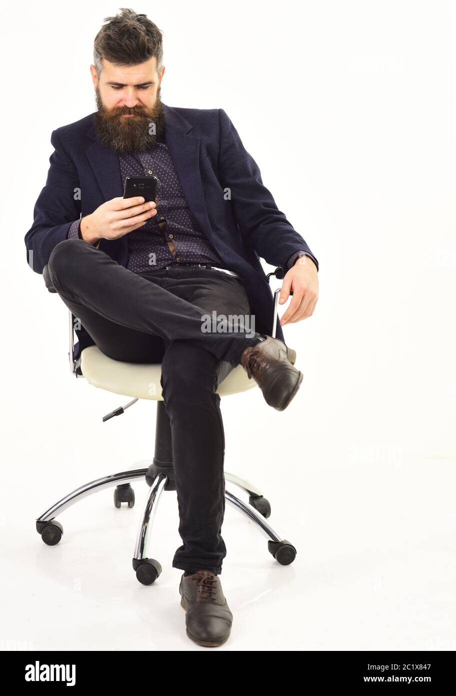 CEO sits in office chair and looks busy. Bearded man looks at smartphone. Businessman with long beard and serious face. Mature man uses mobile phone. Management, business, leadership concept. Stock Photo