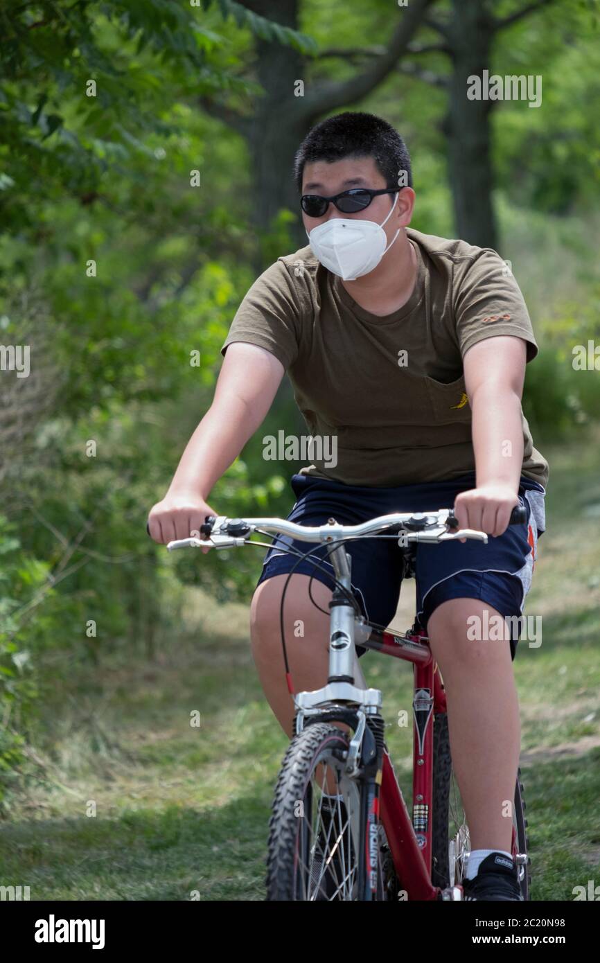 An Asian American young man, likely Korean, bikes on a path near the Cross island Parkway wearing a surgical mask. In Bayside, Queens, NYC. Stock Photo