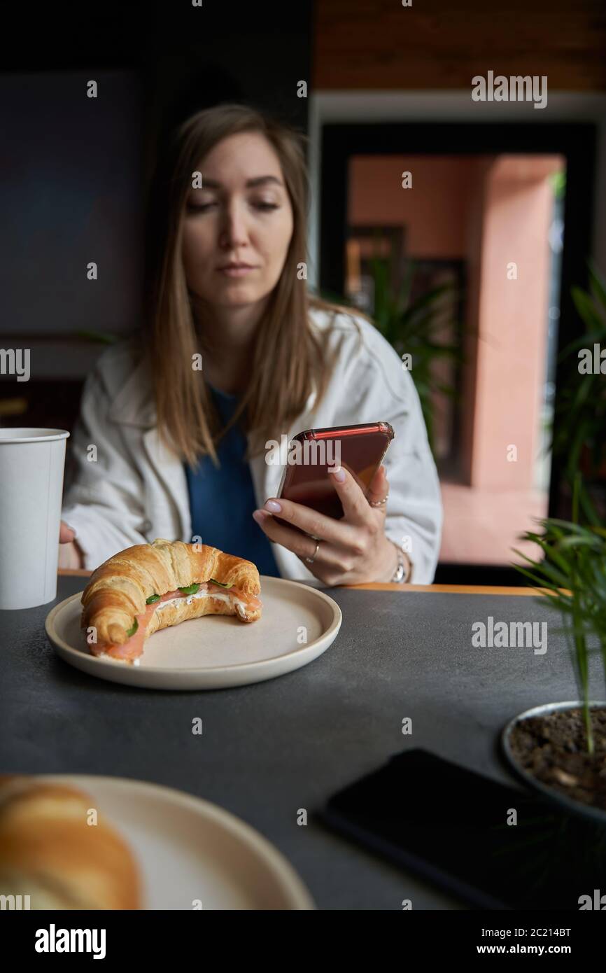 Front view of attractive woman wearing glasses using smartphone, drinking coffee in cafe. Female texting and sharing messages on social media Stock Photo