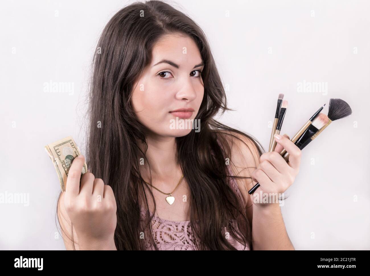 A young girl with a funny expression on her face  while holding money in one hand and make up brushes in the other. Stock Photo