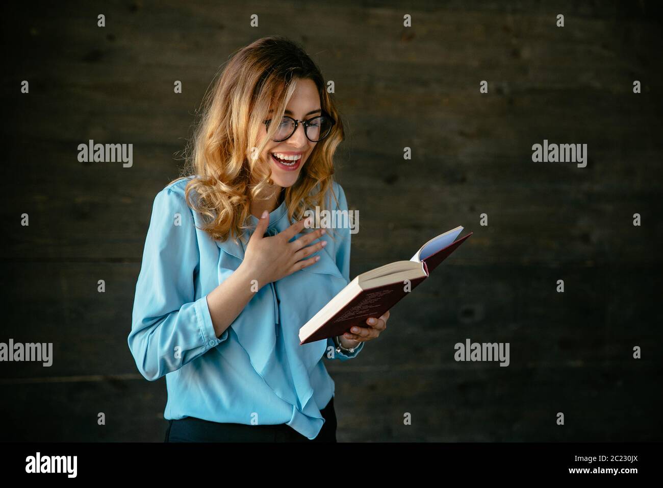Charming young woman bursting out of laugh after reading something funny in book. Dressed in elegant blouse, in eyeglasses. Stock Photo