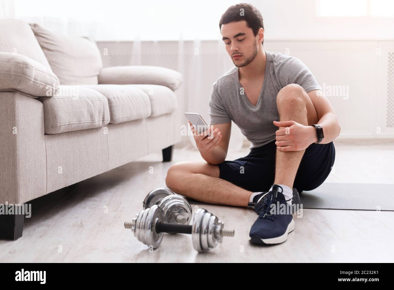 Sports equipment and activities. Man with fitness tracker sitting on floor, looking at smartphone, dumbbells near Stock Photo