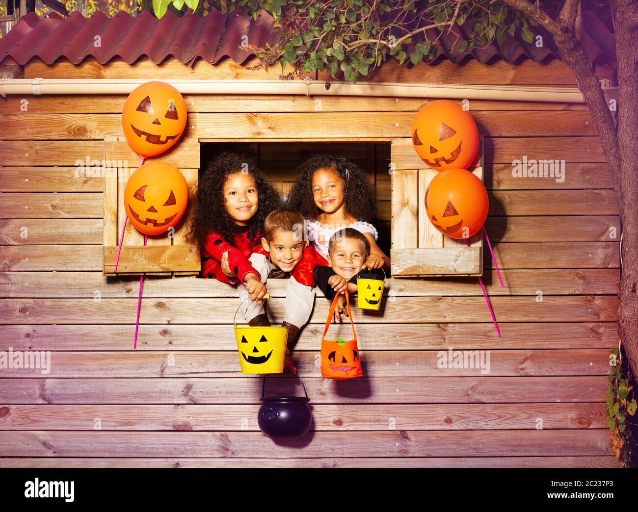Group of kids portrait in Halloween costumes with candy buckets look from the treehouse window decorated using orange balloons Stock Photo