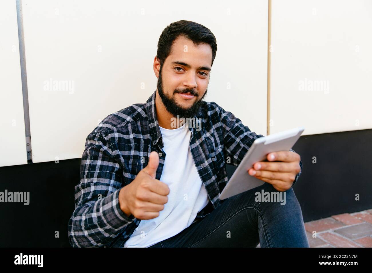 Attractive bearded man showing a thumb up, looking at camera while holding a digital tablet and sitting near the urban wall, outdoors. Stock Photo