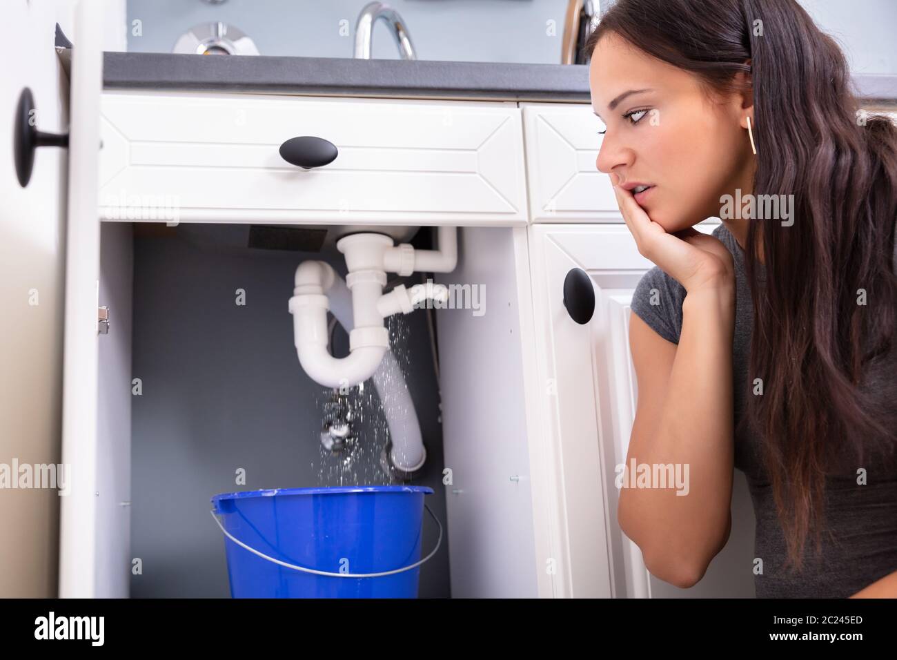 Young Woman Collecting Leakage Water In Bucket At Kitchen Stock Photo