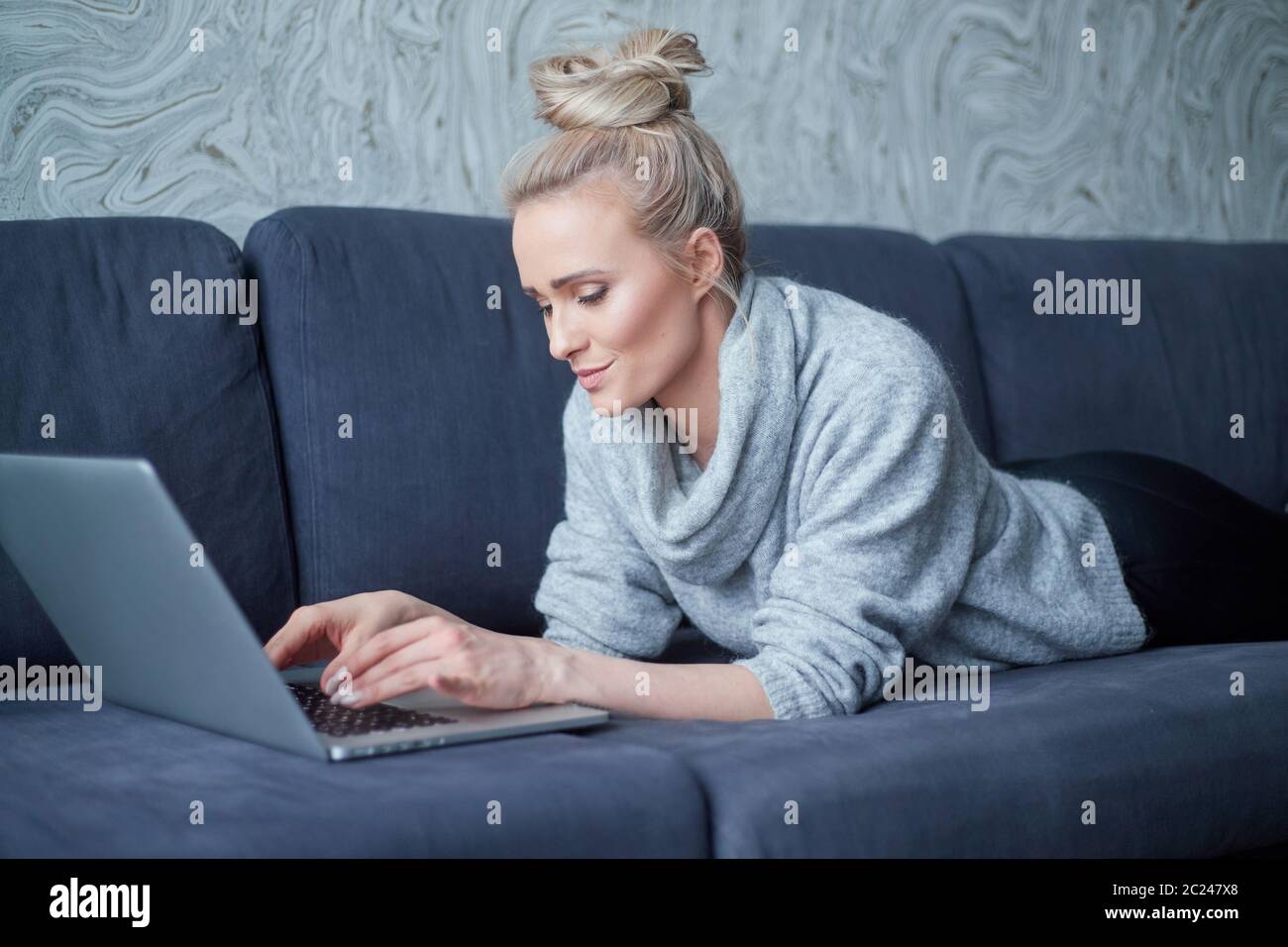 Happy blond woman lying prone on sofa and working on laptop computer Stock Photo