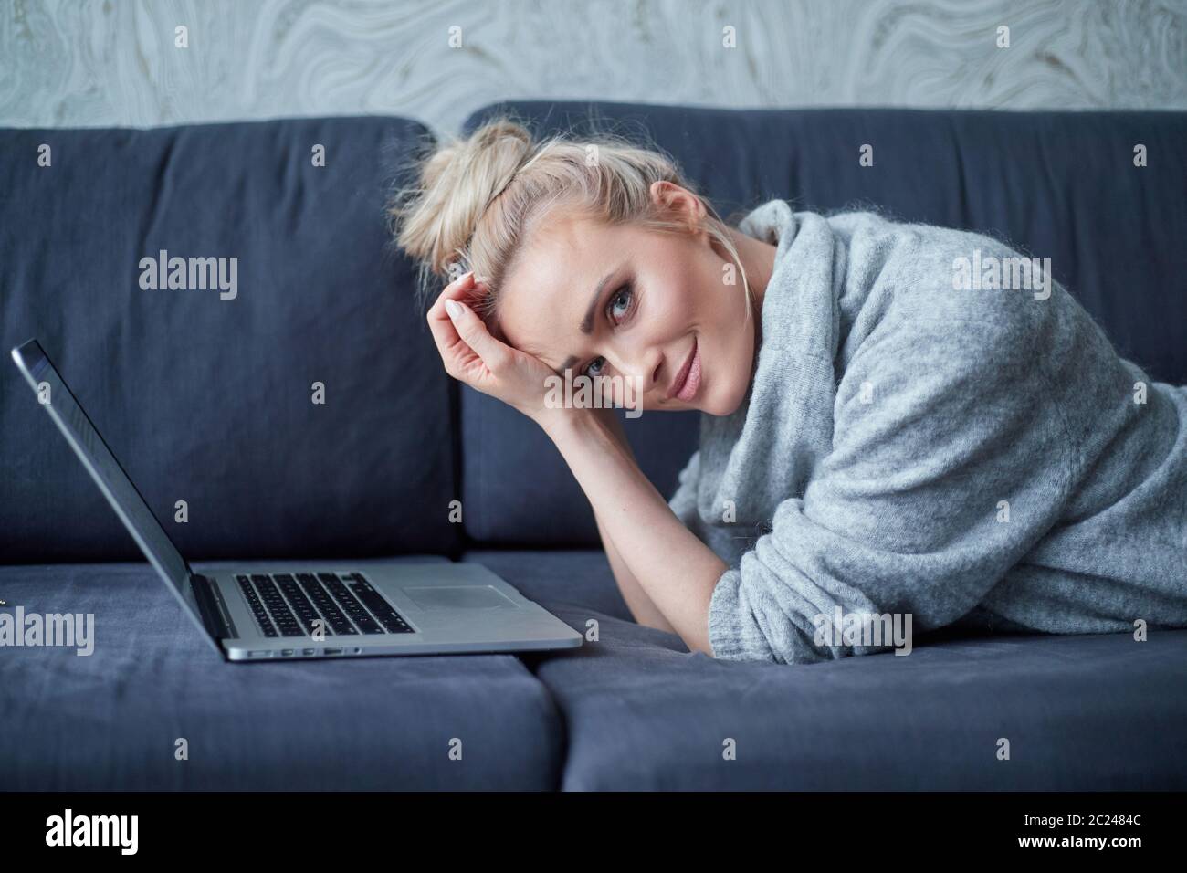 Happy blond woman lying prone on sofa and working on laptop computer Stock Photo