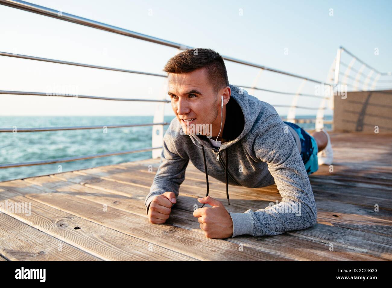Outdoor photo of a purposeful sportive man in headphones, doing exercises, in plank position on elbows. Workout on quay, near the sea. Stock Photo