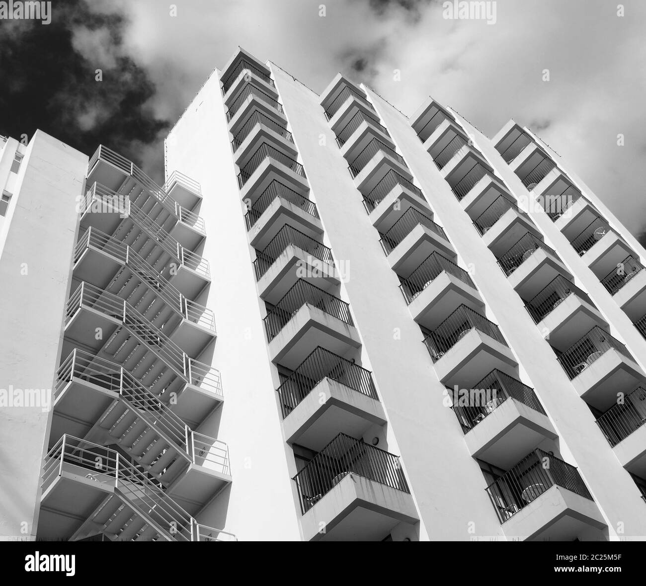 monochrome angled detail view of an old 1960s white concrete apartment building with steps and balconies against sky and clouds Stock Photo