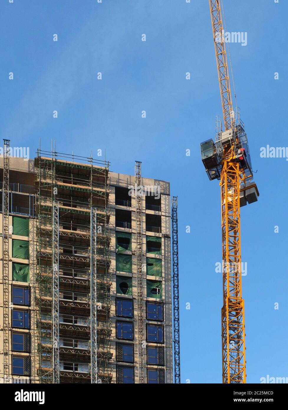 a tall yellow tower crane working on a large hight rise concrete building with scaffolding against a blue sky Stock Photo