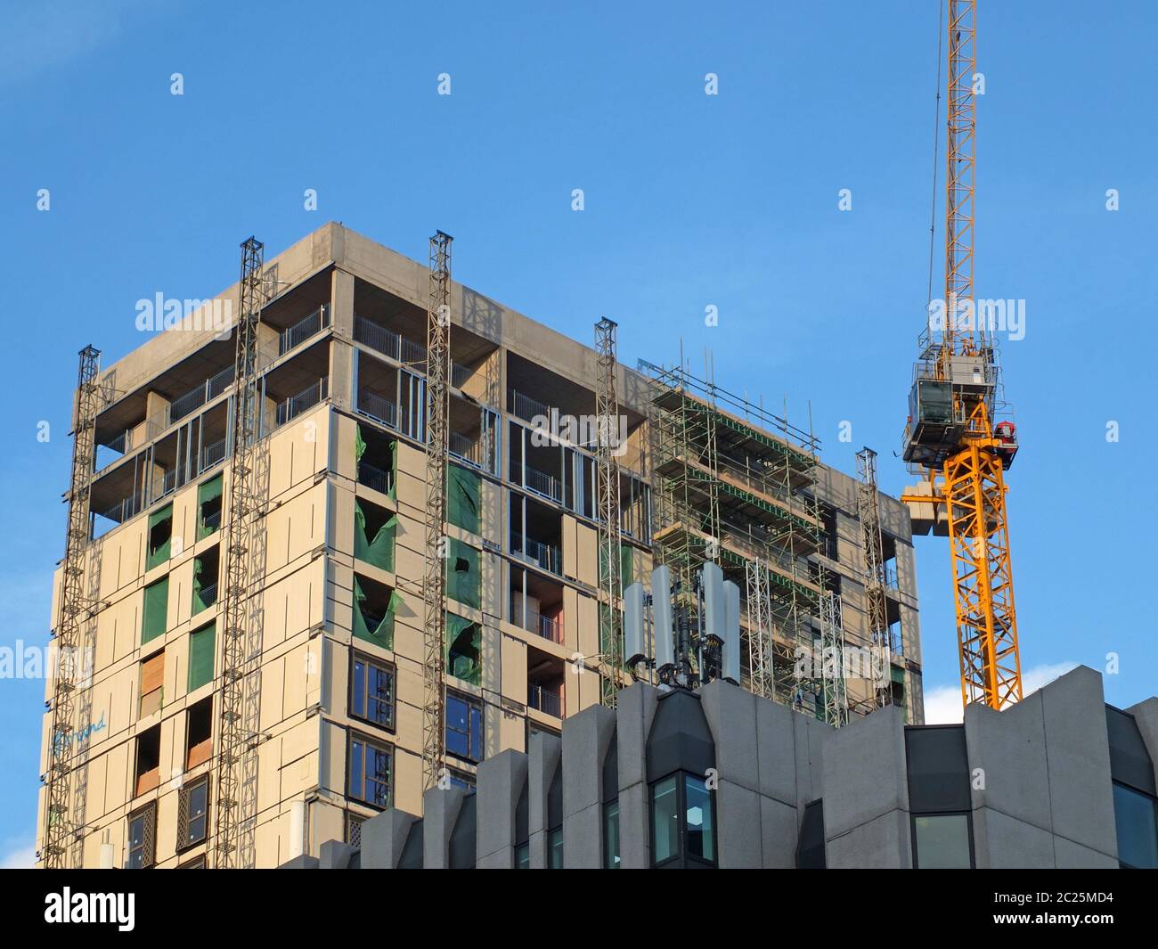 a tall yellow tower crane working on a large hight rise concrete building with scaffolding against a blue sky Stock Photo