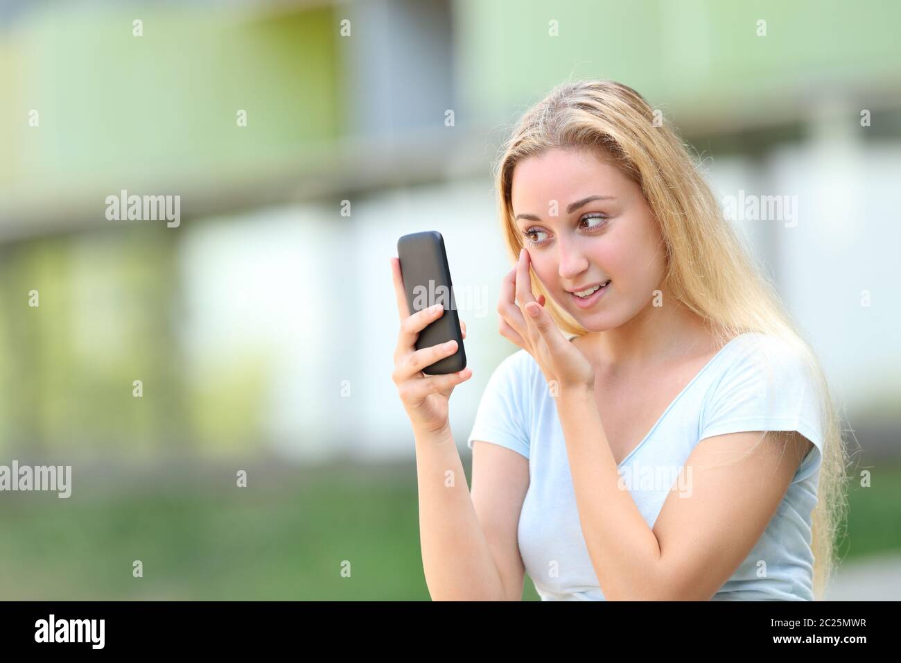 Teenage girl using smart phone as a mirror outdoors in an university campus Stock Photo