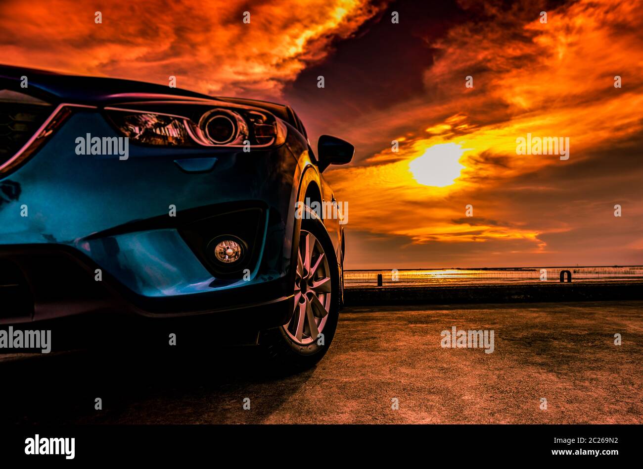 Blue compact SUV car with sport, modern, and luxury design parked on concrete road by the sea at sunset. Front view of beautiful hybrid car. Driving w Stock Photo