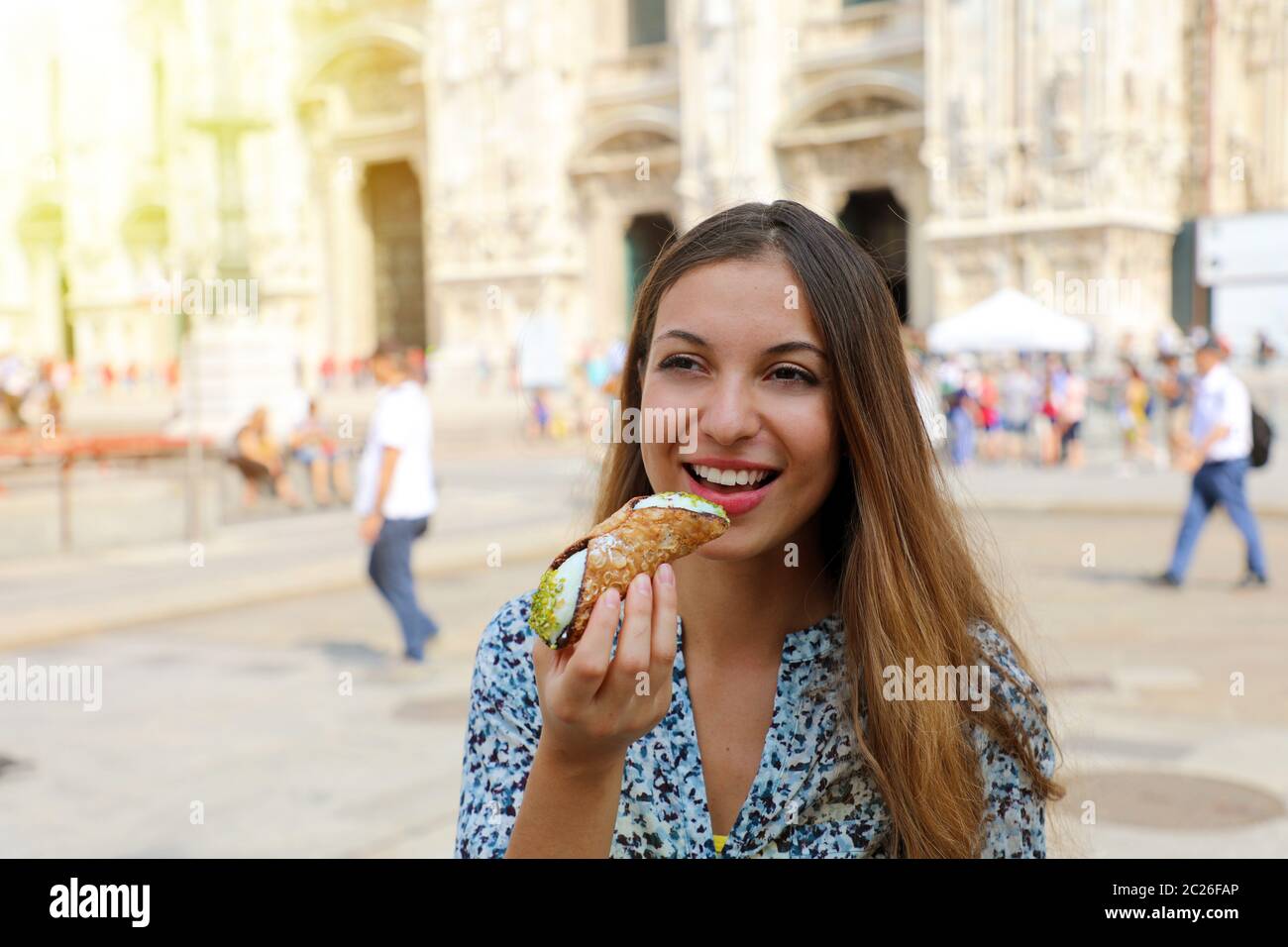 Young female tourist eating cannoli (cannolo siciliano) with Milan Cathedral on the background, Italy Stock Photo