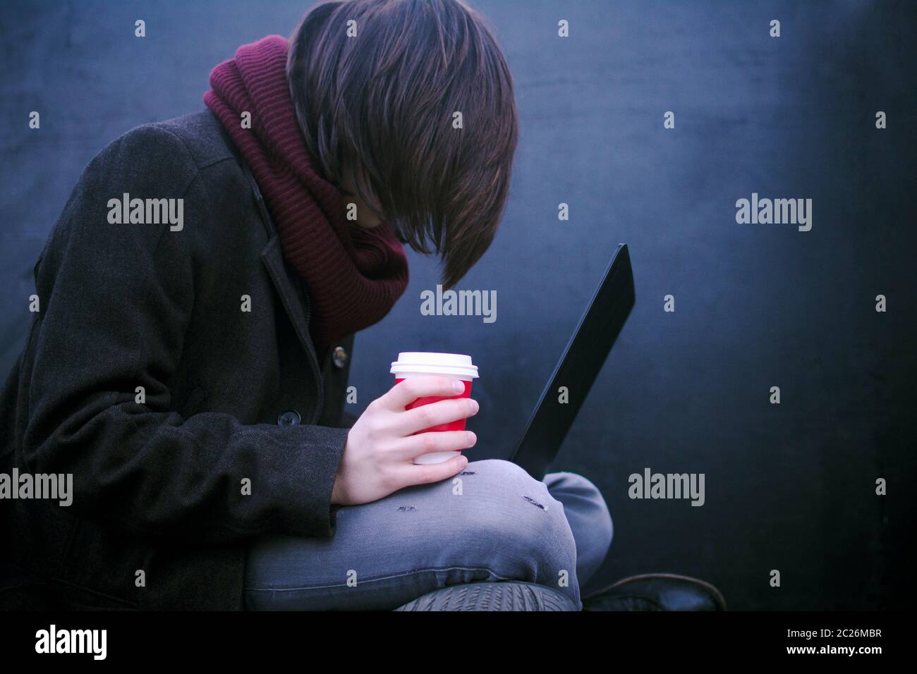 The hacker or freelancer person with coffee in hand is using laptop on dark background. Stock Photo