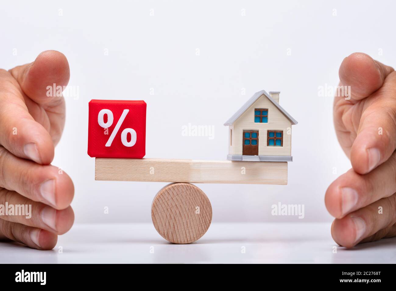 Businessperson's Hand Protecting Balance Between Percentage Red Cubic Block And House Model On Wooden Seesaw Stock Photo