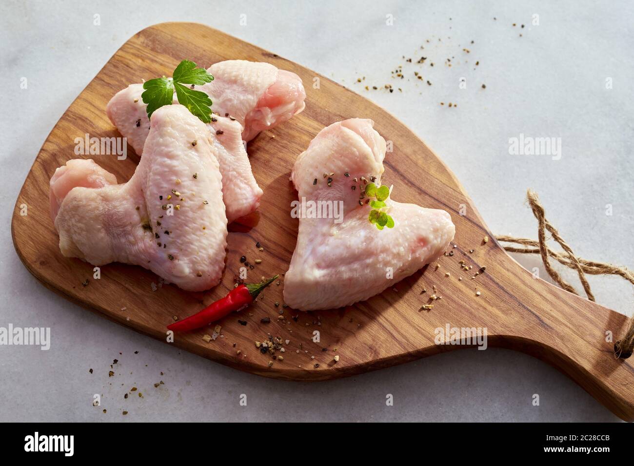 Portions of raw chicken with herbs and spices and red hot chili peppers on a wooden cutting board over a white marble counter in an overhead view Stock Photo