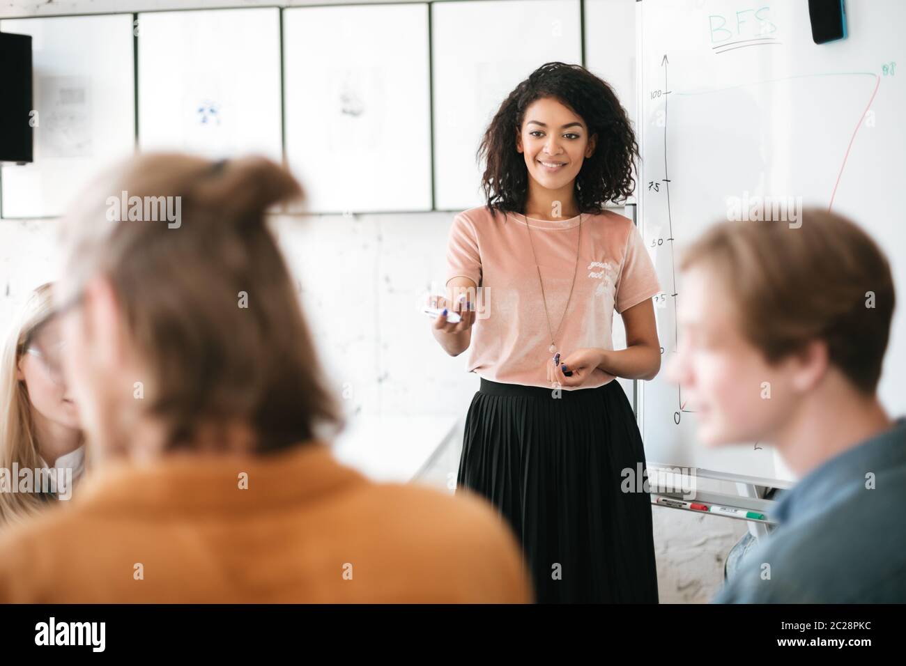 Portrait of beautiful African American lady with dark curly hair standing near board and giving presentation to colleagues in office. Young smiling bu Stock Photo