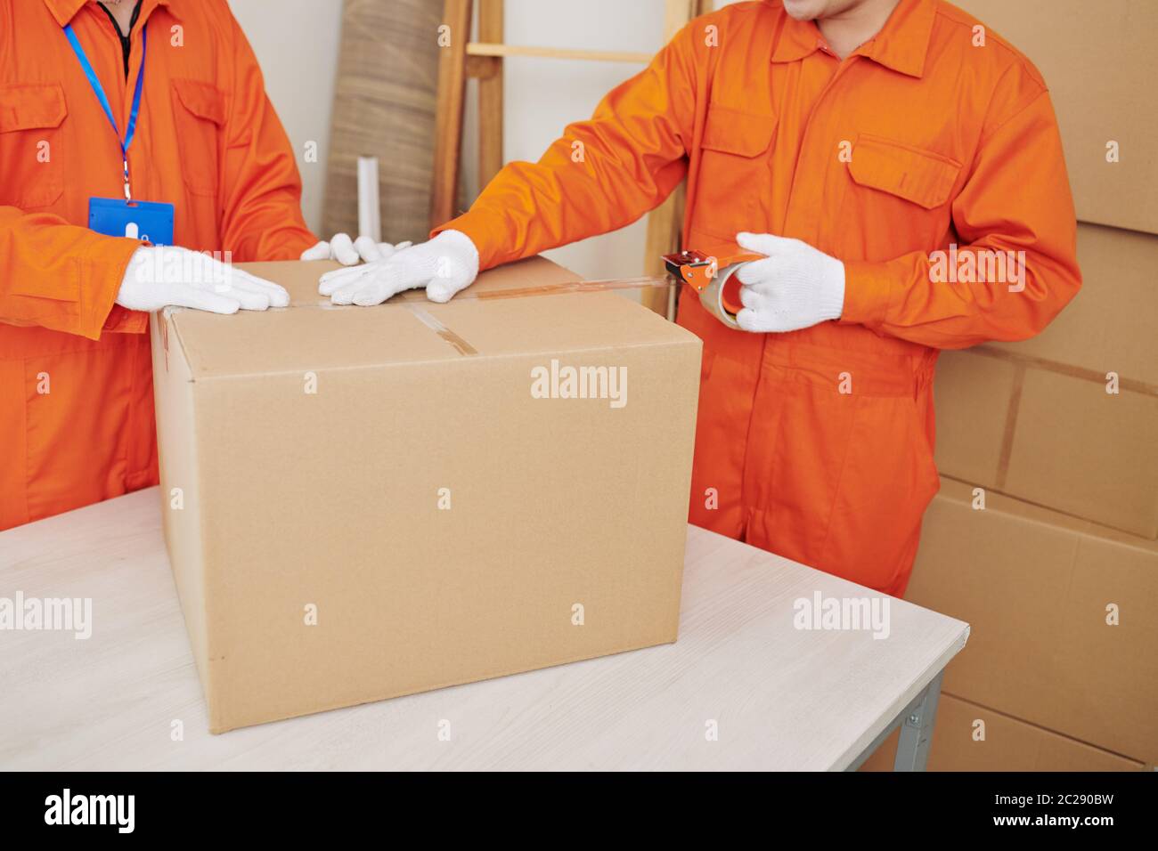 Two unrecognizable house moving service workers wearing bright orange uniform packaging things using boxes and tape Stock Photo