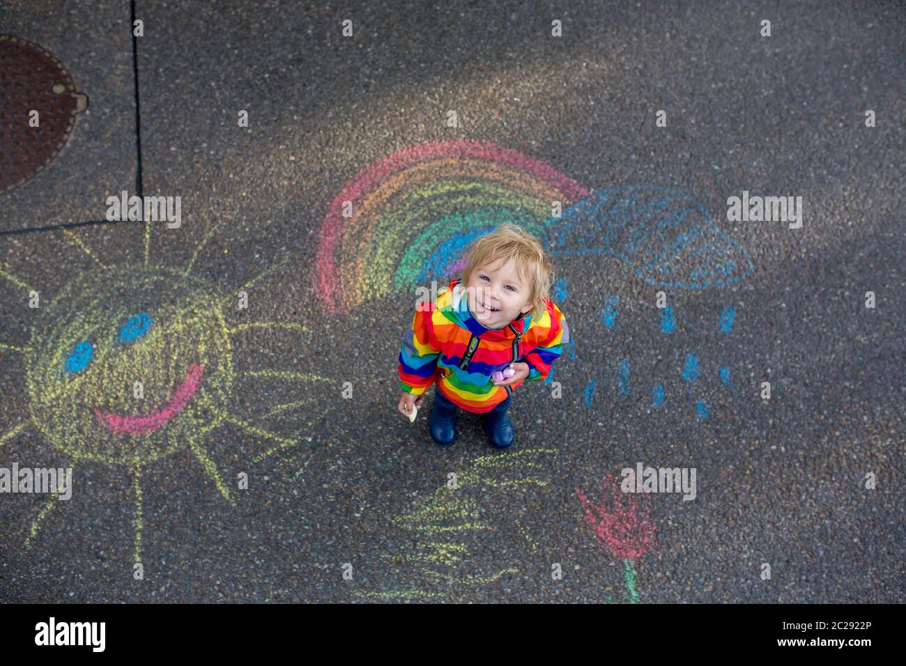 Cute toddler. playing in the rain with chalks, drawing on the asphalt
