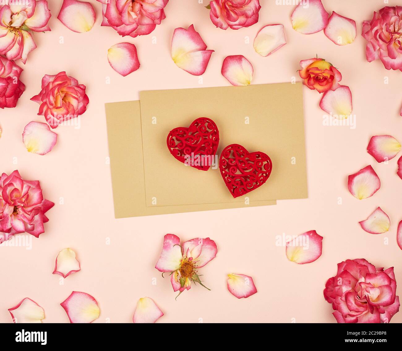 brown paper envelopes and two red carved hearts in the middle of rosebuds, top view, festive backdrop Stock Photo